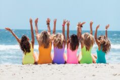 A group of women in colourful swimsuits sitting together on a beach with their arms raised
