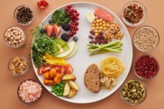 Overhead view of a white plate divided into vegetables, fruit, wholegrains and protein foods, surrounded by small bowls of nuts, spices and pink salt